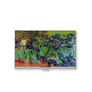 May include: A metal business card holder featuring a print of Vincent van Gogh's "Irises" painting. The artwork depicts blue irises, green foliage, and a yellow iris against a colorful background. The holder has a silver finish and a curved design.