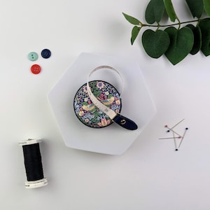 K&ouml;nnte beinhalten: Ein rundes, marineblaues Ma&szlig;band mit Blumen- und Vogelmuster. Das Band erstreckt sich aus einem marineblauen Kunstleder-Etui. Links befinden sich drei Kn&ouml;pfe in Rot, T&uuml;rkis und Marineblau. Eine Spule schwarzen Fadens und Nadeln sind ebenfalls sichtbar.