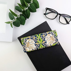 Op de afbeelding: Een brillenkoker met een bloemmotief in cr&egrave;me, groen en geel op een marineblauwe achtergrond. De koker ligt op een zwart boek, met een bril met zwart montuur en groen in de buurt.