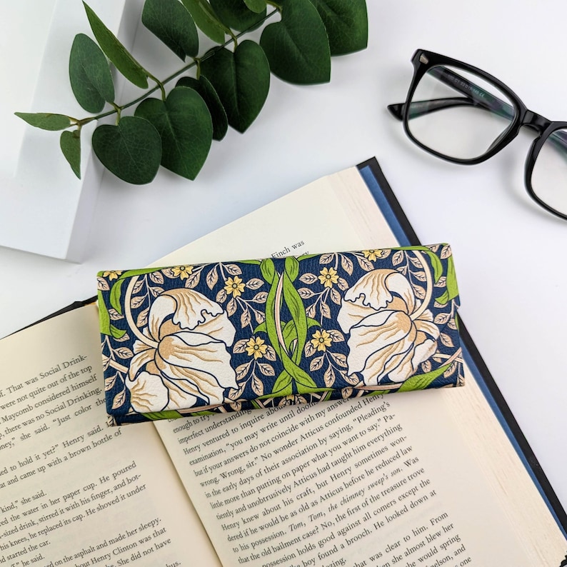Op de afbeelding: Een rechthoekige, open brillenkoker met een bloemenpatroon. De koker heeft een marineblauwe achtergrond met cr&egrave;mekleurige bloemen, groene bladeren en gele accenten. De koker ligt op een open boek.