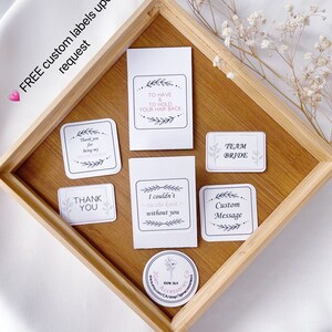 May include: A set of eight white labels with black text and floral designs. The labels include phrases like "Thank you for being my bridesmaid", "To have and to hold your hair back", "Team Bride", "I couldn't tie the knot without you", "Custom Message", and "Thank you".