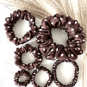 May include: A collection of brown satin scrunchies with white polka dots. The scrunchies vary in size, from small to large, and are arranged on a white surface. The fabric has a slight sheen, and the polka dots are evenly spaced.