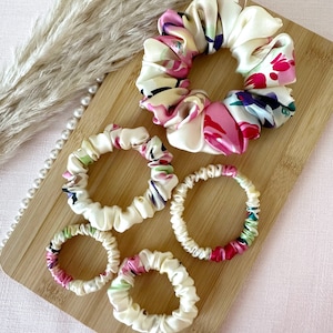 May include: Five colourful floral patterned scrunchies on a wooden surface. The scrunchies are made of soft fabric and are perfect for holding hair in place.