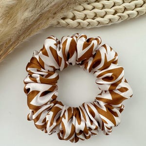 May include: A brown and white patterned scrunchie. The hair accessory features a repeating geometric design in a warm brown color against a white background. The scrunchie is made of a soft, silky material and is gathered to create a voluminous look.