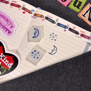 K&ouml;nnte beinhalten: Zwei quadratische, durchscheinend wei&szlig;e Gummistempel mit blauen Tintenmotiven eines Halbmondes mit Gesicht und einer Ansammlung von Sternen. Die Stempel liegen auf einer linierten Notizbuchseite mit einem bunten Buntstift-Rand.