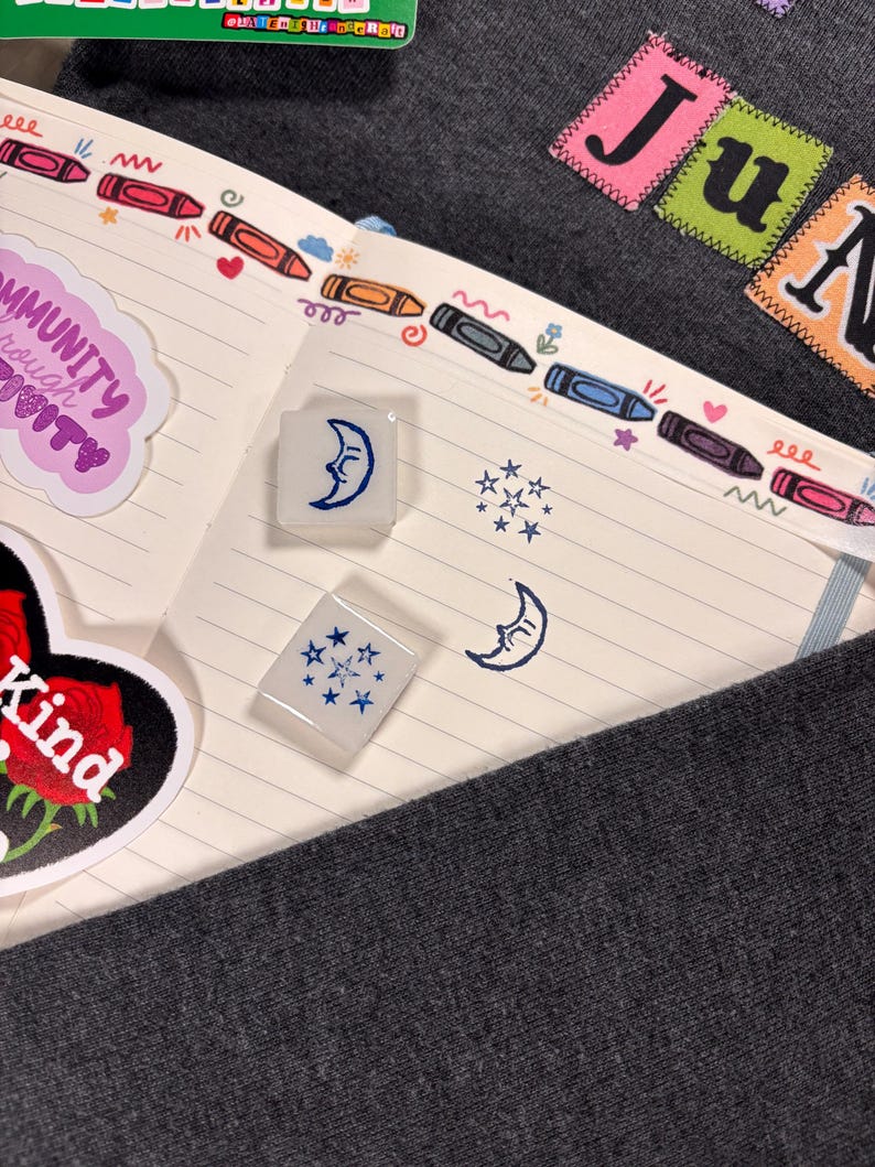 K&ouml;nnte beinhalten: Zwei quadratische, durchscheinend wei&szlig;e Gummistempel mit blauen Tintenmotiven eines Halbmondes mit Gesicht und einer Ansammlung von Sternen. Die Stempel liegen auf einer linierten Notizbuchseite mit einem bunten Buntstift-Rand.