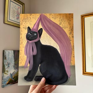 Könnte beinhalten: Eine skurrile Illustration einer schwarzen Katze mit einem rosa Zaubererhut und Schal. Die Katze sitzt und blickt nach oben. Der Hintergrund zeigt ein goldenes Damastmuster. Das Kunstwerk wird von einer Person gehalten.