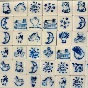 Può includere: Una collezione di piastrelle quadrate bianche con illustrazioni blu. I disegni includono lune, gatti, case, anatre, arcobaleni, orsacchiotti, fiori, stelle e i testi "Love You" e "Handle with Care".