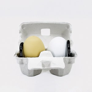 May include: Two silicone eggs in a white cardboard egg carton. One egg is yellow and the other is white.