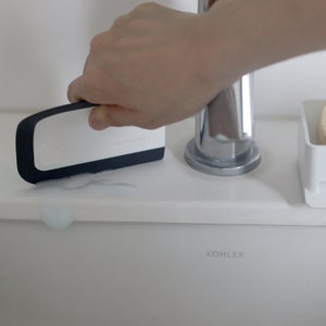May include: A white and black squeegee with the text "unichrome" printed on it. The squeegee is being used to clean a white porcelain toilet bowl with the word "KOHLER" printed on it.
