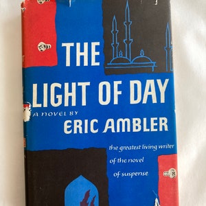 May include: A blue and red book cover with the title "THE LIGHT OF DAY" and the author's name "ERIC AMBLER". The cover also includes the text "A novel by" and "the greatest living writer of the novel of suspense".