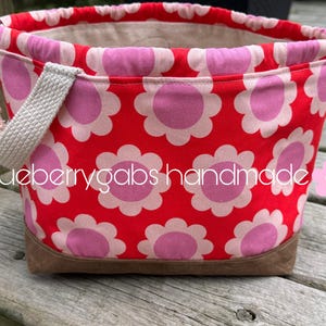May include: A fabric drawstring bag featuring a red background with a repeating pattern of pink flowers with light pink centres. The bag has a beige woven handle, a brown base, and white rope drawstrings. The text blueberrygabs handmade is visible.