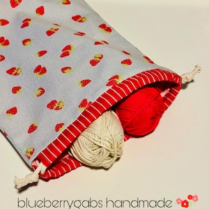 May include: A fabric drawstring bag with a light blue background and a pattern of red strawberries. The bag's interior is red with white stripes, and it contains two balls of yarn, one cream and one red. The text "blueberrygabs handmade" is visible.