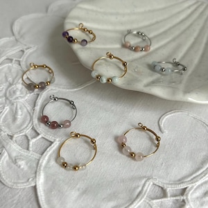 Peut inclure: Plusieurs bagues faites à la main, ornées de perles de pierres précieuses colorées. Les anneaux sont en fil d'or et d'argent, chacun orné de trois ou quatre petites perles rondes de couleurs violettes, roses et blanches. Les bagues sont présentées sur un plat blanc en forme de coquillage.