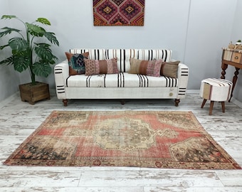 May include: A vintage, hand-woven rug with a red, beige, and brown color scheme. The rug has a distressed, worn look and features a central medallion design. The rug is laid out on a whitewashed wood floor in front of a white sofa with striped cushions.