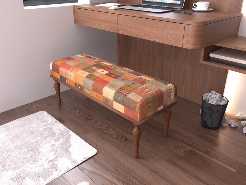a modern, minimalist living room with a wooden desk and a colorful bench. The desk has a laptop and a mouse on it, and there is a trash can nearby. The floor is made of wood, and there is a rug on the floor.