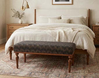 May include: A bedroom scene featuring a bed with a cream-colored headboard and bedding. A patterned bench with wooden legs sits at the foot of the bed. A rug with a faded pattern and a wooden nightstand are also visible.
