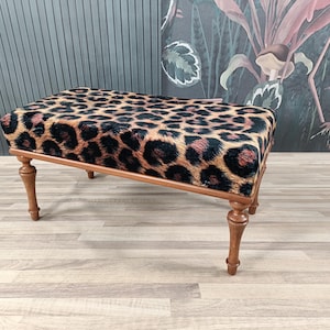 May include: A leopard print upholstered bench with wooden legs. The bench is on a light wood floor.
