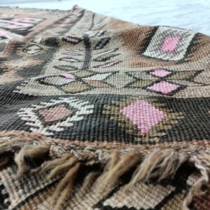 May include: A vintage rug with a geometric pattern in shades of brown, pink, and white. The rug has a fringe border and is woven with a thick, textured yarn.