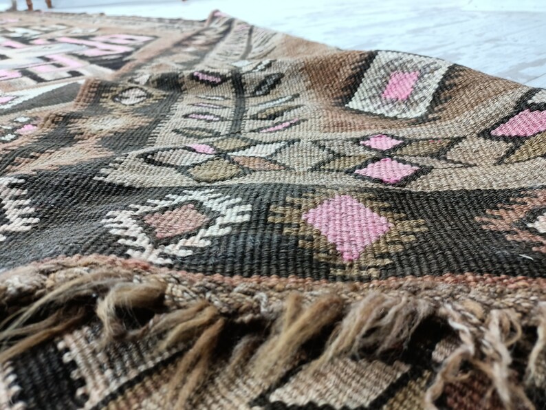 May include: A vintage rug with a geometric pattern in shades of brown, pink, and white. The rug has a fringe border and is woven with a thick, textured yarn.
