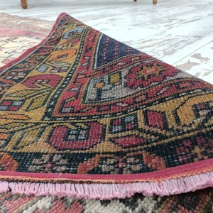 May include: A close-up of a colorful, hand-woven rug with intricate patterns in shades of red, yellow, blue, and black. The rug has a pink fringe border.