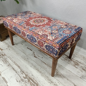 May include: A rectangular bench with a patterned cushion in shades of red, blue, and beige. The cushion features a central starburst design. The bench has four wooden legs.