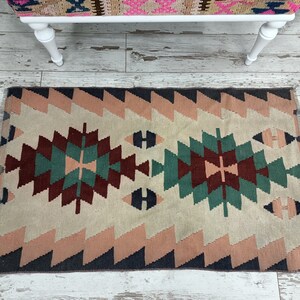 K&ouml;nnte beinhalten: Ein rechteckiger Teppich mit geometrischem Muster. Der Teppich hat ein zentrales Design in Burgunder-, T&uuml;rkis- und Cremet&ouml;nen, das von einem Zickzack-Rand in Rosa und Marineblau umrahmt wird. Das Design des Teppichs ist symmetrisch und hat einen Fransen.