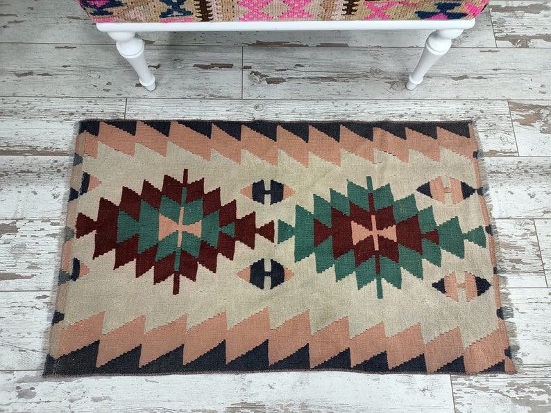 K&ouml;nnte beinhalten: Ein rechteckiger Teppich mit geometrischem Muster. Der Teppich hat ein zentrales Design in Burgunder-, T&uuml;rkis- und Cremet&ouml;nen, das von einem Zickzack-Rand in Rosa und Marineblau umrahmt wird. Das Design des Teppichs ist symmetrisch und hat einen Fransen.