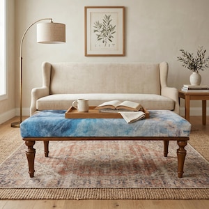 May include: A living room scene with a blue and white patterned ottoman with a wooden frame and turned legs. A wooden tray holds a mug, book, and folded cloth. A beige sofa, floor lamp, and framed botanical print complete the decor.