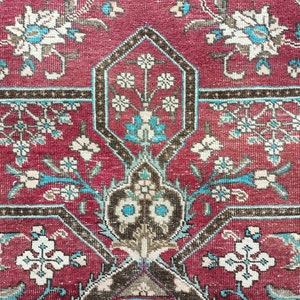 May include: A close-up of a red rug with a geometric pattern of white and blue flowers. The rug has a brown border.