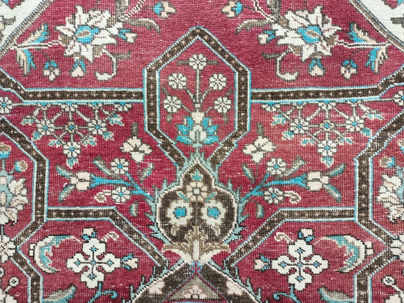 May include: A close-up of a red rug with a geometric pattern of white and blue flowers. The rug has a brown border.