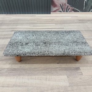 May include: A small, rectangular, black and white patterned ottoman with wooden legs. The ottoman is sitting on a wooden floor.