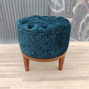 Puede incluir: Un otomano tapizado de color azul turquesa con patas de madera. El otomano está sobre un suelo de madera clara.