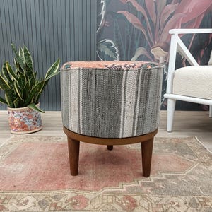 Puede incluir: Un otomán redondo y tapizado con un cuerpo de tela a rayas grises y blancas. La parte superior presenta una tela estampada en tonos rosas, verdes y marrones. Descansa sobre una base de madera marrón oscuro con tres patas cortas, mostrando texturas y colores.