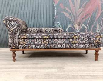 May include: A patterned chaise lounge with a dark brown wood frame and legs. The chaise lounge is upholstered in a dark brown fabric with a geometric pattern in shades of pink, yellow, and blue.