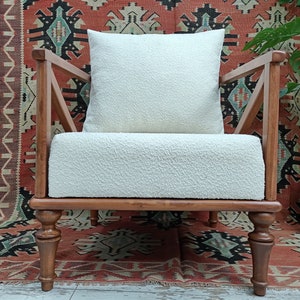May include: A white armchair with a white fluffy cushion, wooden arms, and turned wooden legs. The chair is in front of a red, black, and white patterned rug.