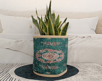 Puede incluir: Un macetero de tela con estampado verde y rosa con una planta serpiente en su interior. El macetero está sobre una superficie rayada.