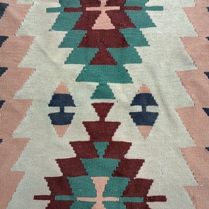 K&ouml;nnte beinhalten: Gewebter Teppich mit geometrischem Muster. Der Teppich hat ein zentrales Design in Beige-, T&uuml;rkis-, Burgunder- und Marineblaut&ouml;nen. Die R&auml;nder sind dunkelgrau, und das Gesamtdesign ist symmetrisch.
