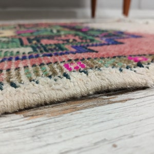 Peut inclure: Gros plan sur un tapis color&eacute; avec une frange blanche. Le tapis pr&eacute;sente un motif g&eacute;om&eacute;trique avec des nuances de rose, de vert, de bleu et de marron.