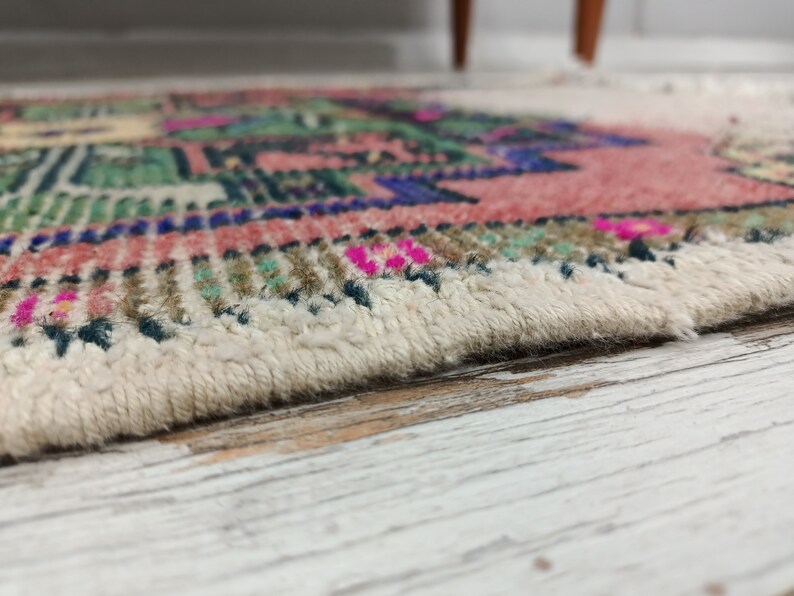 Peut inclure: Gros plan sur un tapis color&eacute; avec une frange blanche. Le tapis pr&eacute;sente un motif g&eacute;om&eacute;trique avec des nuances de rose, de vert, de bleu et de marron.