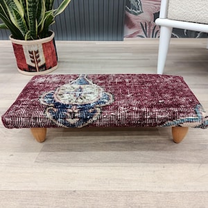 Puede incluir: Un otomán rectangular burdeos con un diseño estampado en azul y blanco. El otomán tiene cuatro patas cortas de madera clara. La textura es similar a la de una alfombra. Mide aproximadamente 60 cm de largo y 30 cm de ancho.