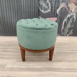 Pode incluir: Um pufe redondo e tufado em um tom verde suave. O pufe tem uma base e pernas de madeira. O design é simples e elegante, adequado para vários estilos de interiores. O pufe tem aproximadamente 46 cm de diâmetro e 41 cm de altura.