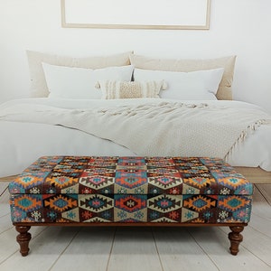May include: A colourful patterned storage ottoman with a wooden frame and four turned legs. The ottoman is in front of a bed with white bedding and a throw blanket.