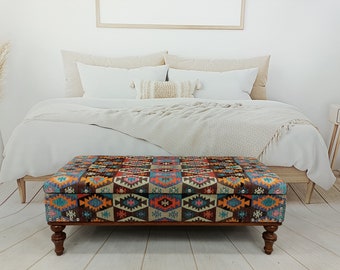 Op de afbeelding: Een kleurrijke, gepatroneerde opbergottoman met een houten frame en vier gedraaide poten. De ottoman staat voor een bed met witte beddengoed en een deken.