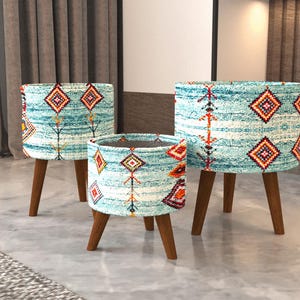 May include: Three cylindrical planters with three wooden legs each, featuring a turquoise and white fabric exterior with a repeating geometric pattern in orange, red, and black. The planters vary in height and width, standing on tapered brown legs.