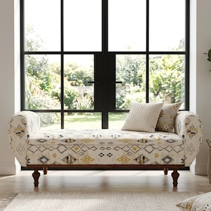 May include: A patterned bench in cream, yellow, and grey, with curved arms and wooden legs. Two cushions rest on the bench. The bench is positioned in front of a large window, offering a view of the garden.
