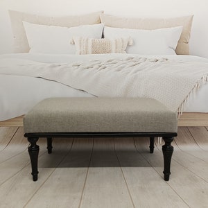 May include: A light grey upholstered ottoman with black turned legs. The ottoman is in front of a bed with white bedding and a white throw blanket.