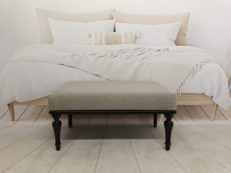 May include: A light grey upholstered ottoman with black turned legs. The ottoman is in front of a bed with white bedding and a white throw blanket.