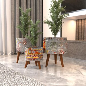 May include: Three cylindrical planters with a gray, red, and yellow patterned fabric exterior, each supported by three tapered wooden legs. Each planter holds a green leafy plant with thin, pointed foliage, creating a tiered display.