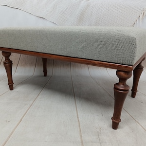 May include: A light grey upholstered bench with a wooden frame and turned legs. The bench has a simple, elegant design and would be a great addition to any home.
